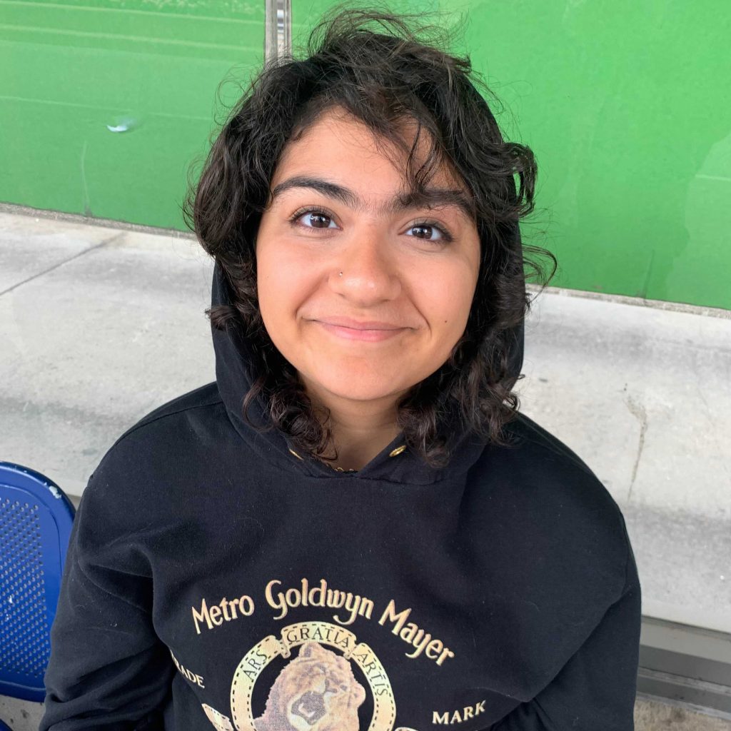 Layla, a brown teenager, smiles on a bus stop bench. She is wearing a Metro Goldwyn-Meyer hoodie with the hood on and curls peeking out.