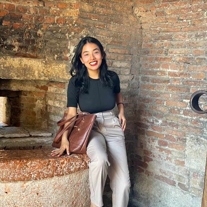 A petite brown woman smiling in red lipstick and business casual clothing leans against a large brown brick fireplace.