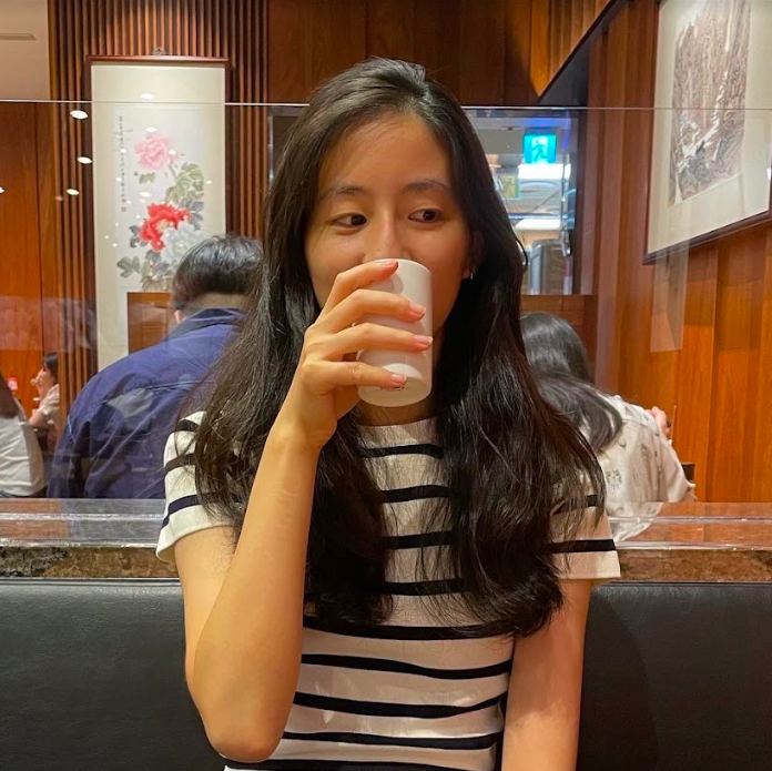 An East Asian girl wearing a black-and-white striped shirt sits in a restaurant booth. She stares off to the side while holding a cup to her mouth.